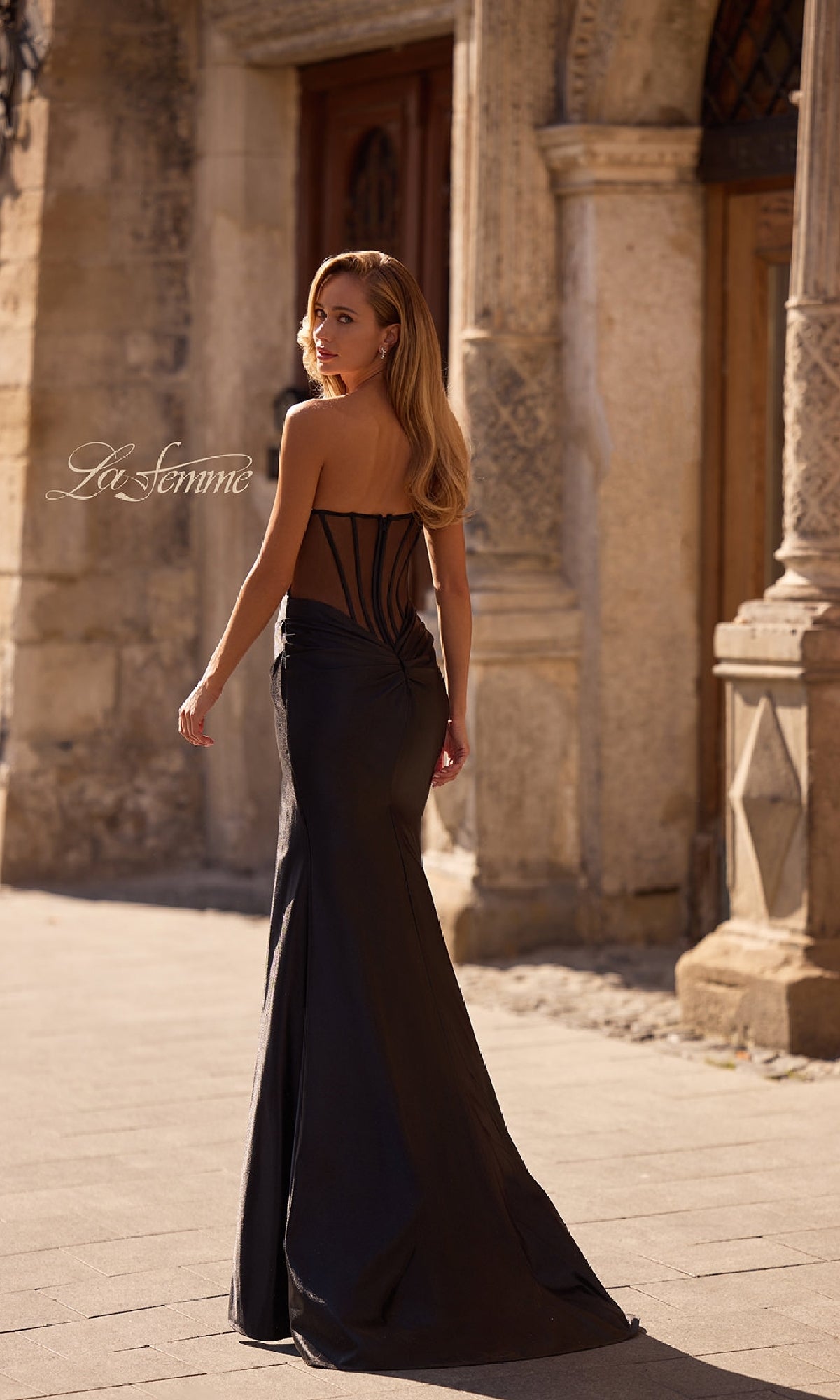 Wearing the Sheer-Back Strapless Prom Gown: La Femme 32649, a woman walks along a sunlit stone pathway beside an old building.